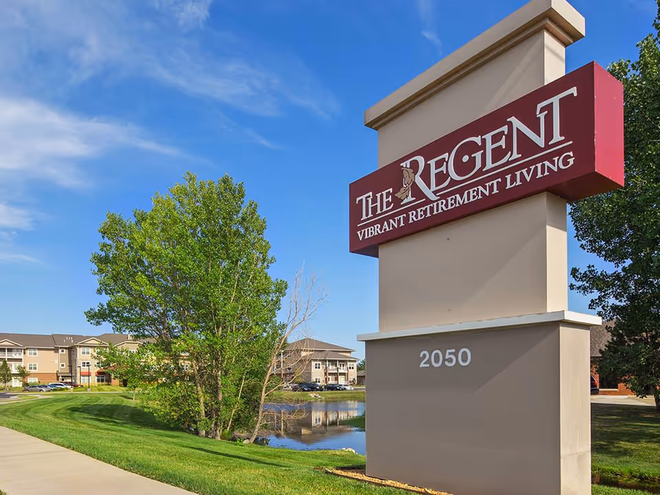 Freestanding sign reading "The Regent Vibrant Retirement Living" with the number 2050 beside a pond, trees, and apartment buildings under a blue sky.