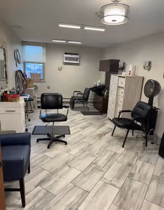 Interior view of a senior living facility room set up as a hair salon with multiple black salon chairs, a hair dryer chair, a cabinet, and a window with an air conditioning unit. The floor is tiled with light-colored tiles, and the walls are painted light gray.