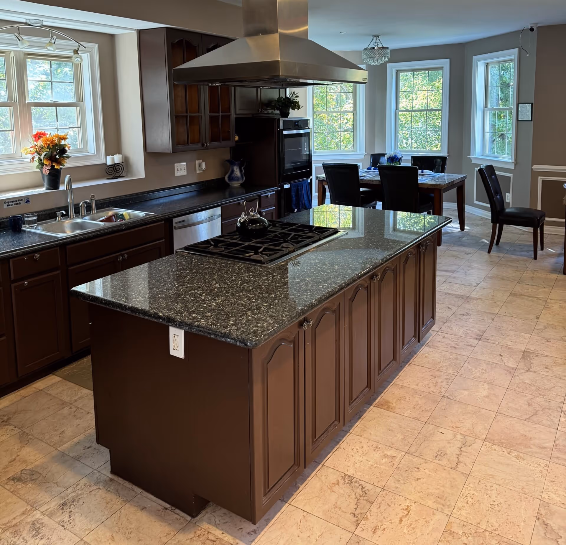 Modern kitchen with dark wood cabinets, granite island, stainless range, and a dining area by windows.