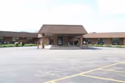 Front exterior view of a single-story building with a covered entrance and a large paved parking lot in front. The building has a brown roof and brick walls with some greenery along the base.
