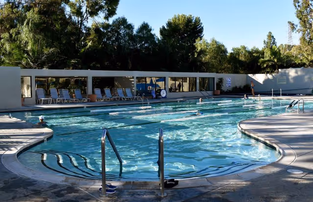 Outdoor swimming pool with several swimmers in the water and lounge chairs lined up along the poolside. Trees and greenery surround the area, and there is a building with large windows in the background.