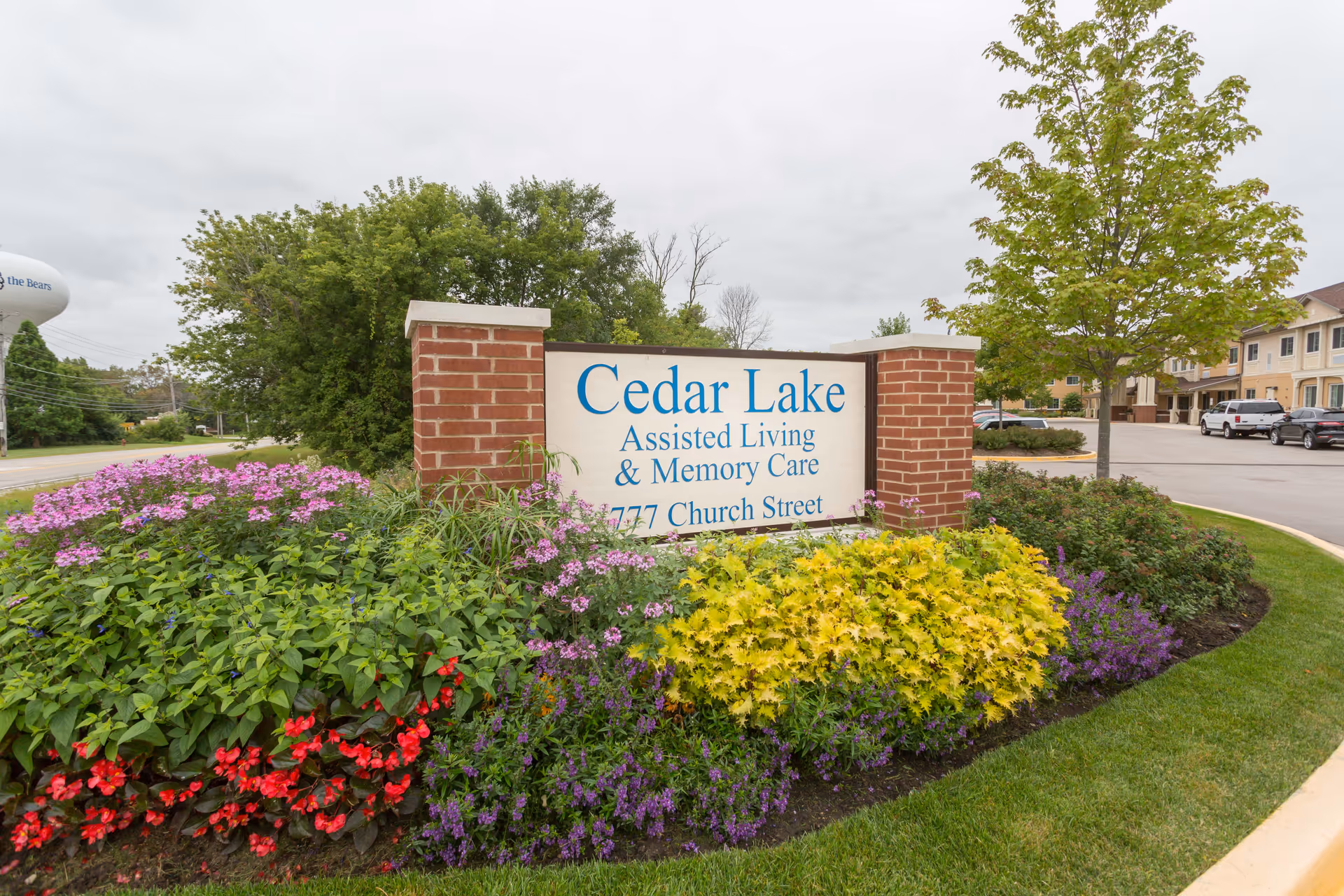 Outdoor view of the entrance sign for Cedar Lake Assisted Living and Memory Care, surrounded by colorful flowers and greenery, with a parking lot and building visible in the background under a cloudy sky.