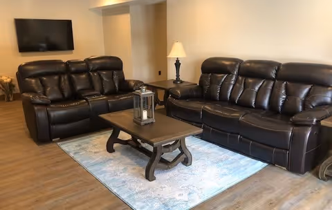 A cozy living room with two dark leather sofas facing a wooden coffee table on a pale blue rug, a wall-mounted TV, and a floor lamp.