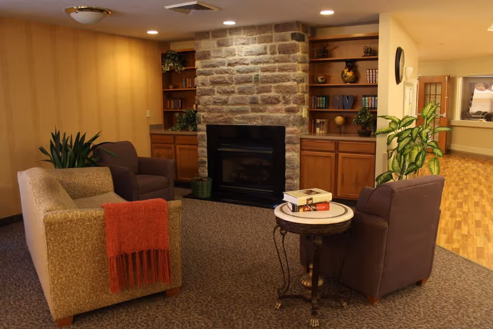A cozy sitting area in a senior living facility with a stone fireplace in the center, flanked by wooden built-in shelves filled with books and decorative items. The seating includes a beige patterned sofa with a red throw blanket and two brown armchairs. A small round table with books on top is placed between the armchairs. There are plants on either side of the fireplace and the room has warm lighting and carpeted flooring. A hallway with wooden flooring and windows is visible in the background.