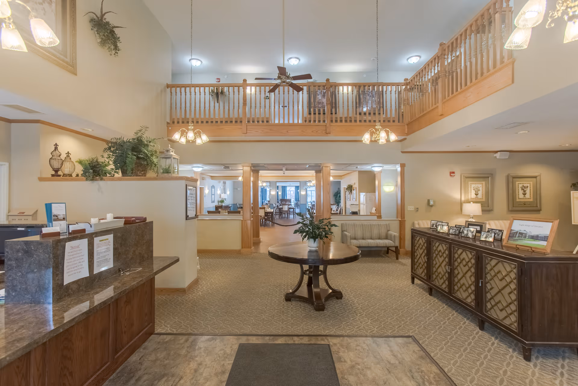 Interior view of a senior living facility lobby with a reception desk on the left, a round wooden table with a plant in the center, and a wooden railing balcony above. The area is well-lit with ceiling lights and hanging lamps, and there are chairs and framed pictures on a cabinet to the right.