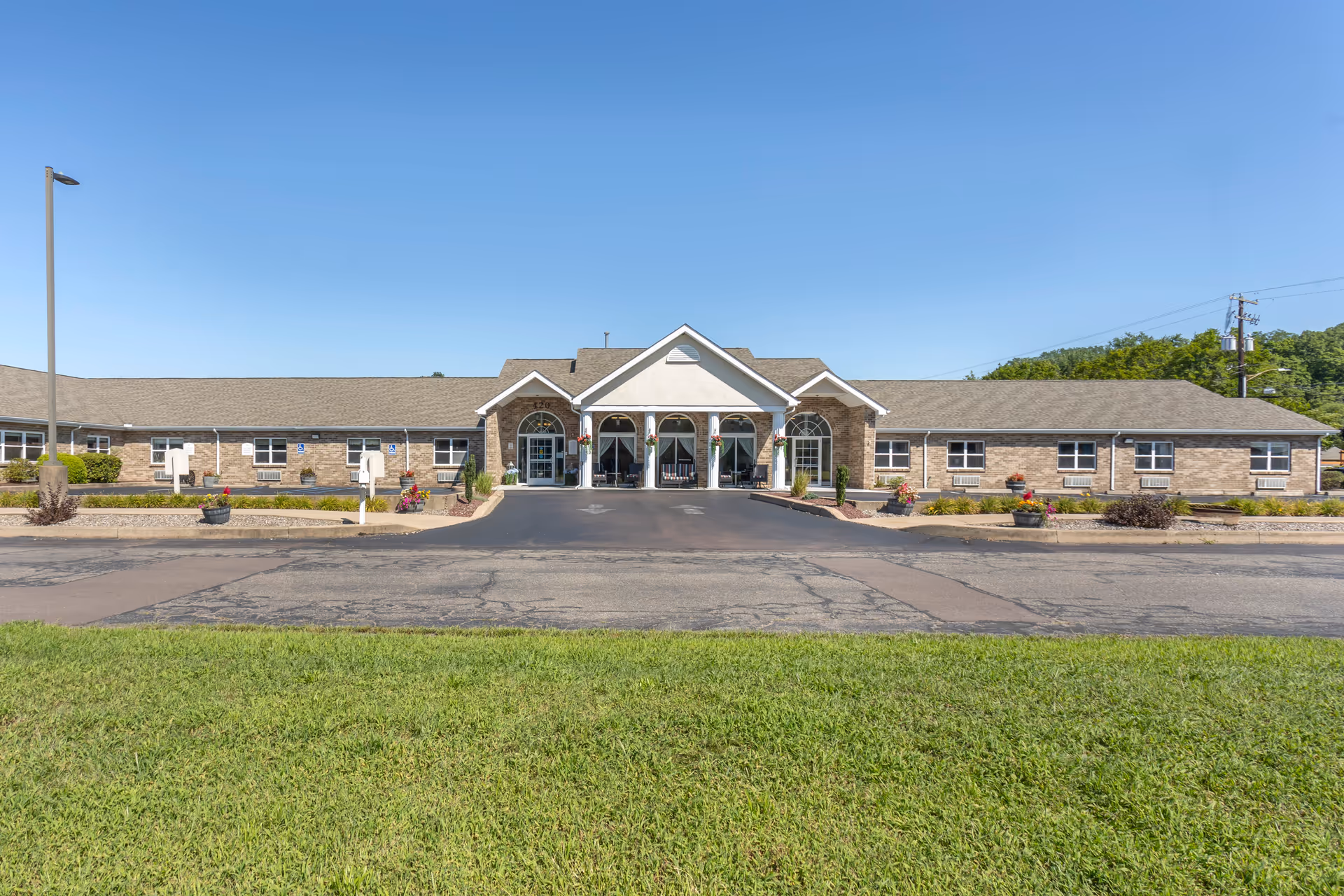 Front exterior view of a single-story senior living facility building with a central entrance featuring a covered portico, surrounded by landscaped areas with flowers and shrubs, under a clear blue sky.