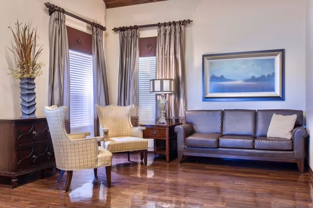 A cozy living room with a brown leather sofa, two beige patterned armchairs, a wooden side table with a lamp, a framed landscape painting on the wall, and two windows with curtains. There is also a decorative vase with dried plants on a wooden cabinet.