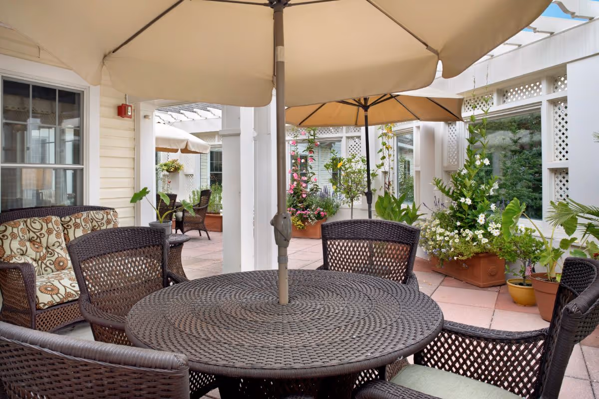 Outdoor patio area with a round wicker table and matching chairs under large beige umbrellas. There are potted plants and flowers around the patio, with a cushioned wicker loveseat visible on the left side. The area is surrounded by white walls with windows and lattice details.