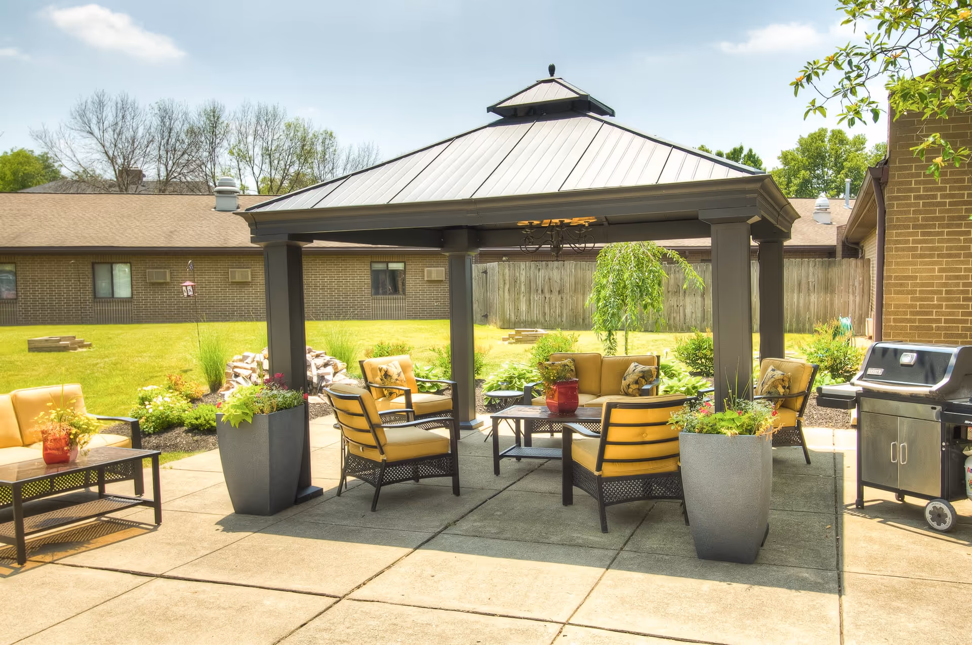 Outdoor patio area at Mayfair Village Retirement Center featuring a gazebo with cushioned seating including chairs and a sofa, a coffee table with a red vase, potted plants, and a barbecue grill on a sunny day with green grass and trees in the background.
