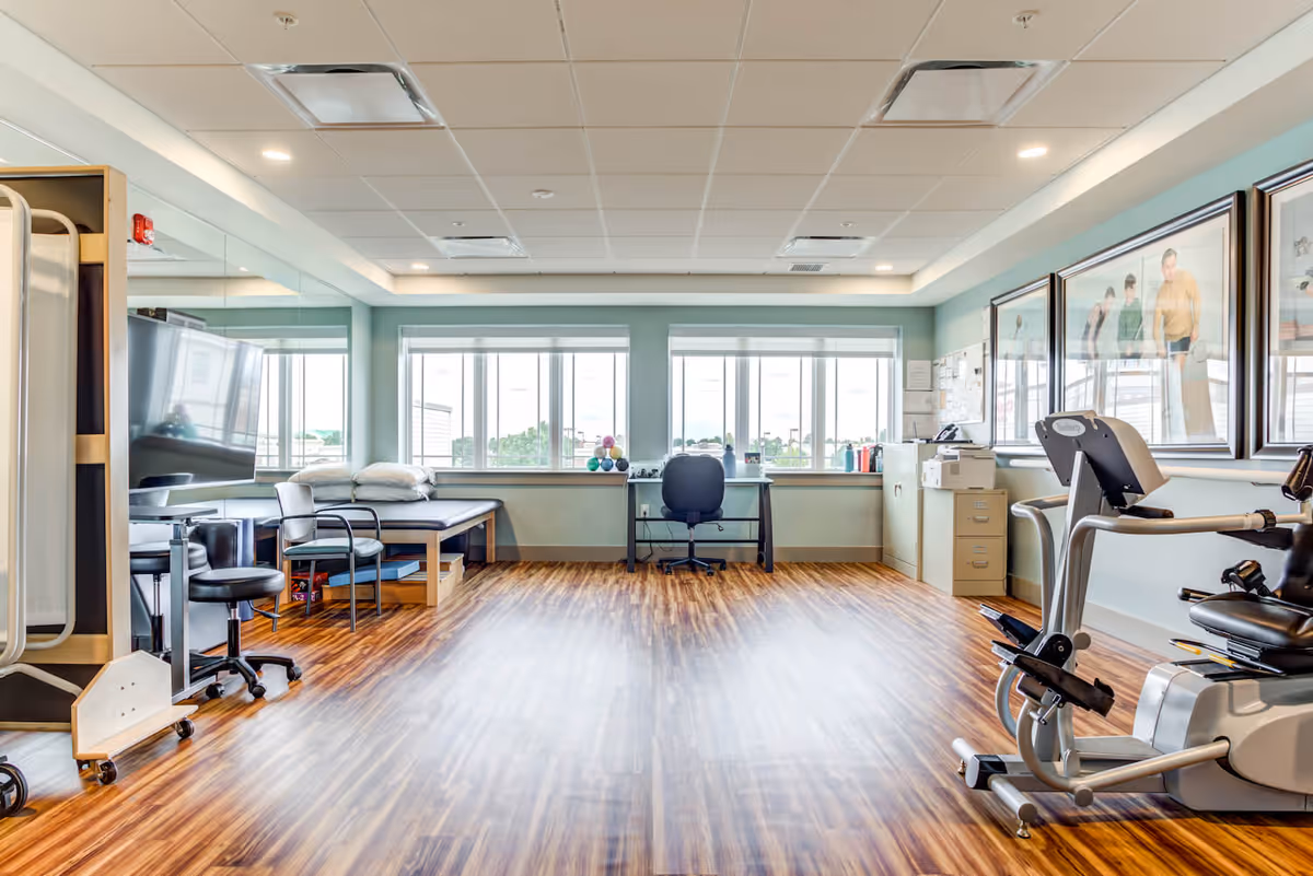 A bright and spacious therapy or exercise room with wood flooring, large windows letting in natural light, a stationary exercise bike, a therapy table with pillows, chairs, a desk with a chair, filing cabinets, and framed pictures on the wall.