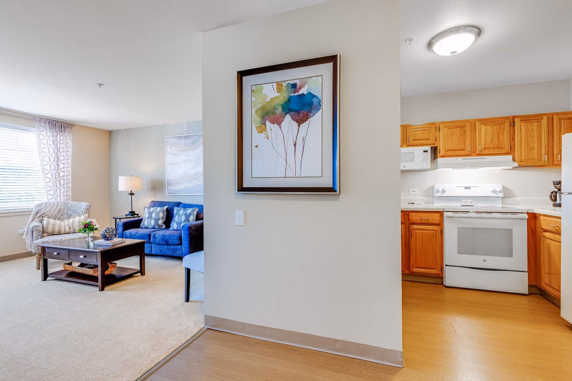 Interior view of a senior living facility showing a cozy living room with a blue sofa, armchair, coffee table, and a lamp next to a large window with patterned curtains. Adjacent to the living room is a kitchen area with wooden cabinets, a white stove, microwave, and refrigerator. A colorful abstract painting is hung on the wall between the living room and kitchen.