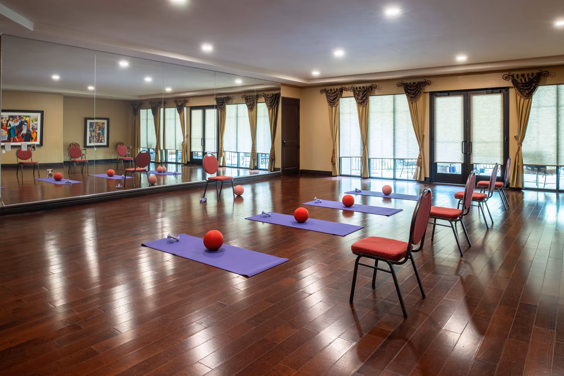A spacious exercise room with wooden floors and large mirrors on one wall. The room has several purple yoga mats each with a red exercise ball and small dumbbells placed on them. Red cushioned chairs are arranged near the mats. Large windows with beige curtains and blinds allow natural light into the room.