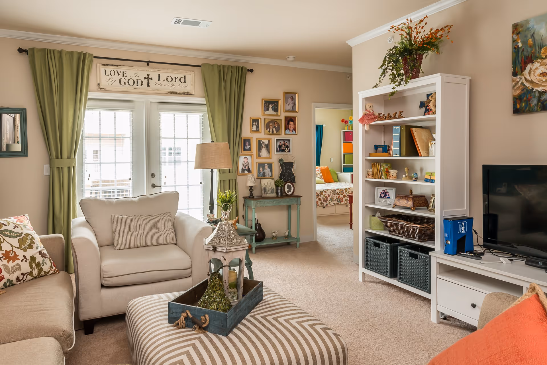 A cozy living room with beige walls and carpet, featuring a beige armchair and sofa with floral and striped cushions. A striped ottoman with a decorative tray and lantern sits in the center. Green curtains frame glass doors letting in natural light. A white bookshelf filled with books and decorative items stands next to a TV on a white media console. A small green side table with framed photos and a lamp is against the wall, and a doorway leads to a bedroom with colorful bedding. A sign above the doors reads 'Love the Lord with all thy heart.'