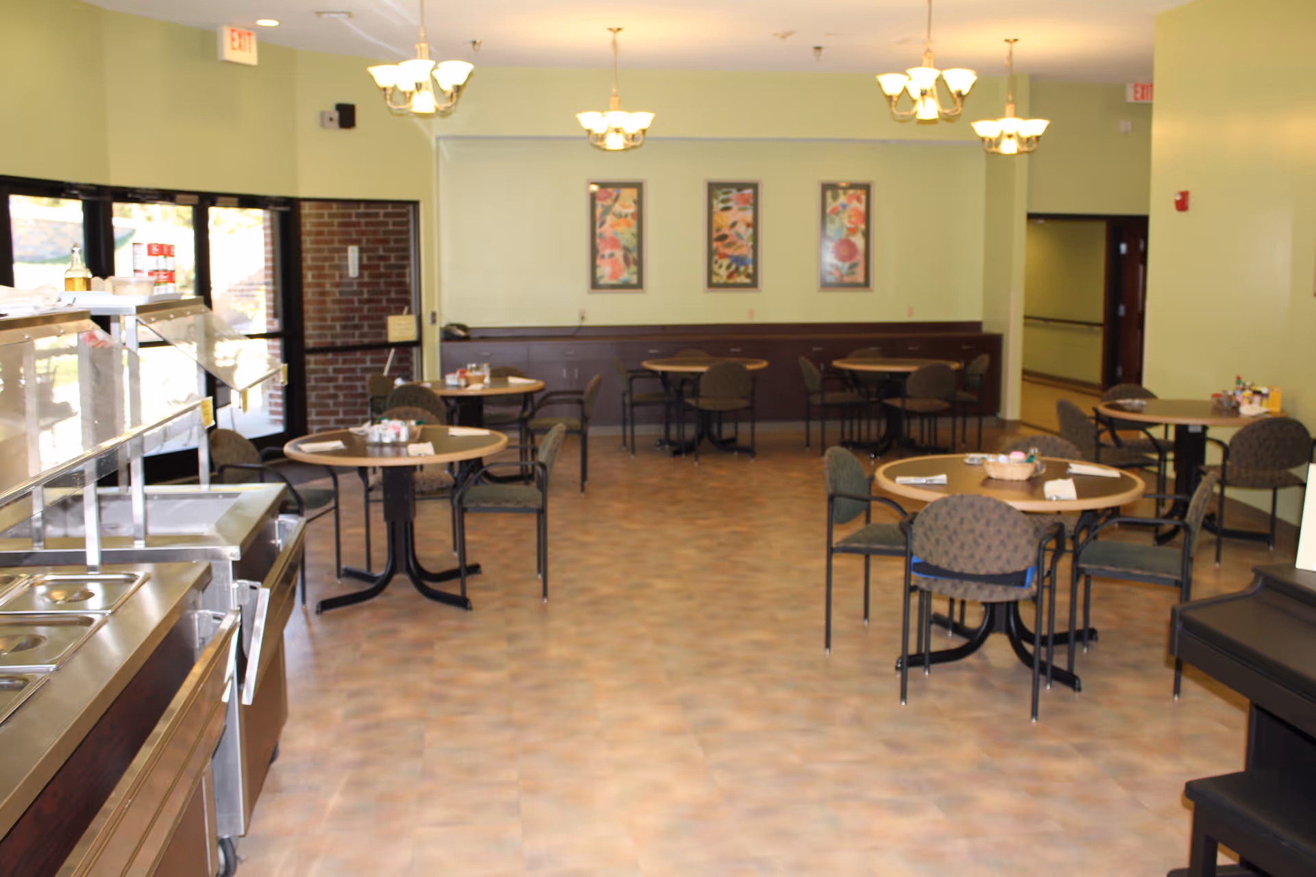 Well-lit dining room with round tables and chairs, a serving counter on the left, and framed artwork on the back wall.