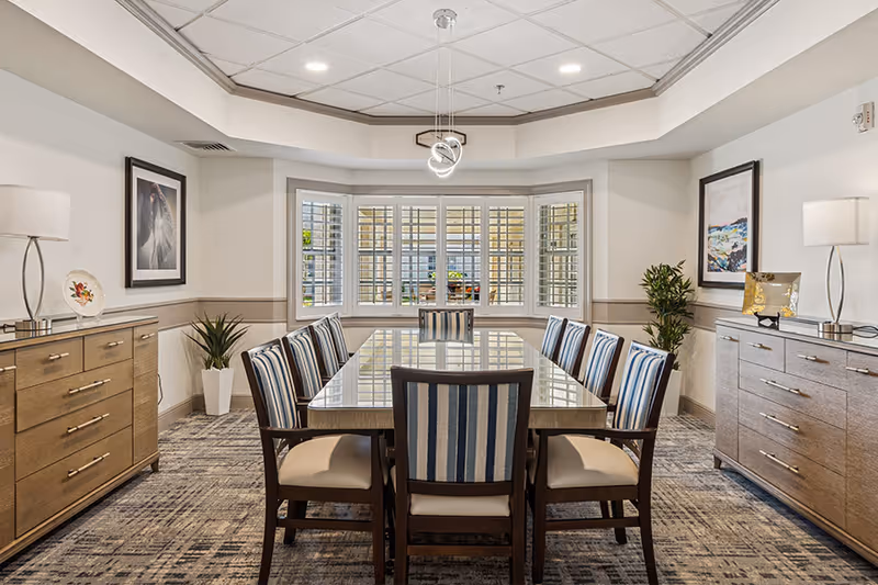 A well-lit dining room with a large rectangular table surrounded by ten chairs with striped upholstery. The room features two wooden sideboards on either side, each with a lamp and decorative items. A large bay window with white shutters is at the far end, and the ceiling has recessed lighting and a modern pendant light fixture.