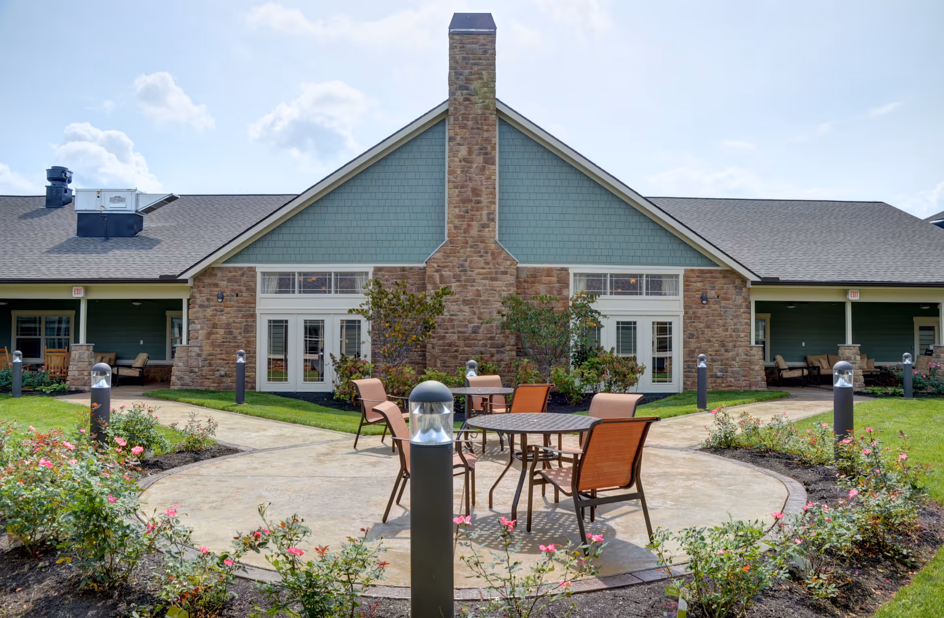Outdoor patio area with round tables and chairs surrounded by flower beds and garden lights, in front of a building with stone and green siding and large windows.