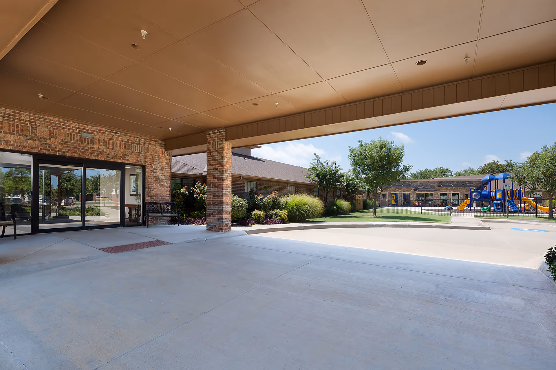 Covered entrance area of a brick building with glass doors leading inside. Outside, there is a paved driveway and a fenced playground with colorful play structures, surrounded by grass and trees under a clear blue sky.