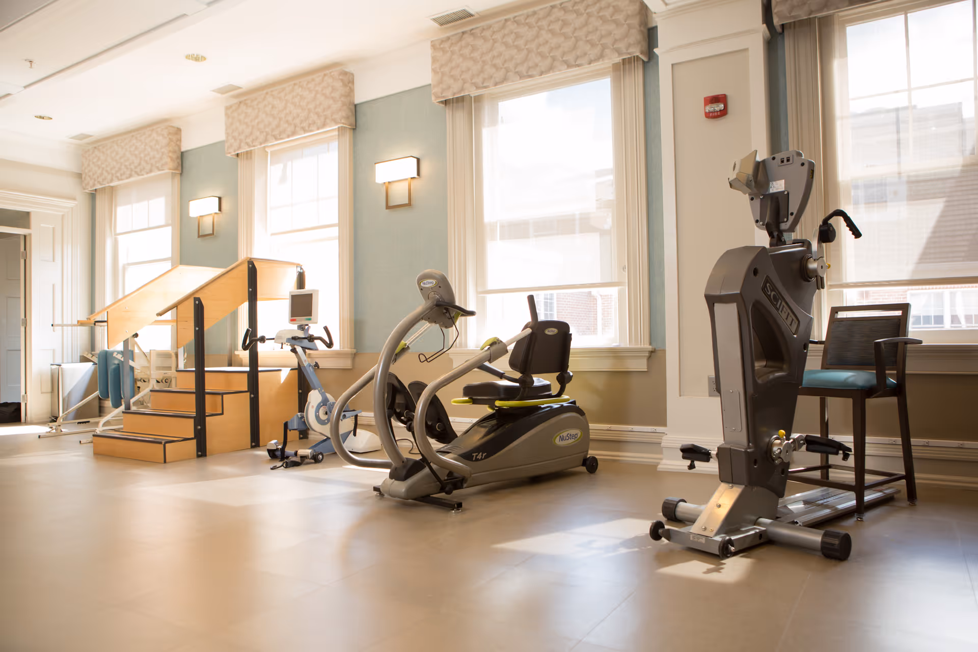 A bright exercise room with large windows covered by light-colored blinds. The room contains various exercise equipment including a recumbent bike, a seated elliptical machine, a stationary bike, and a set of wooden stairs with handrails for physical therapy or rehabilitation. There is also a chair placed near the equipment. The walls are painted light blue with white trim and the floor is a light beige color.