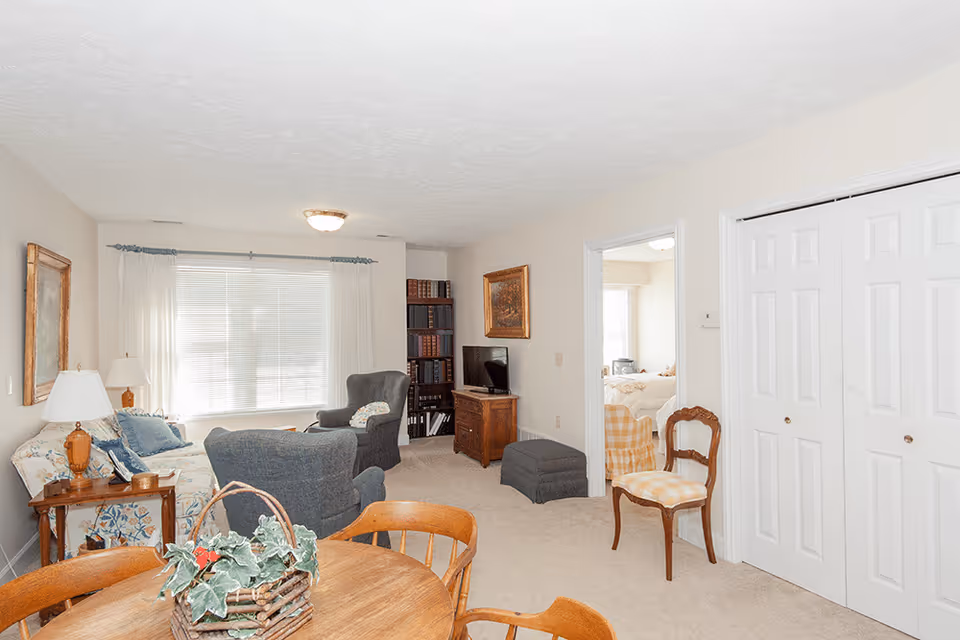 A bright living room with beige walls and carpet, featuring a wooden round table with chairs in the foreground, a floral-patterned sofa with cushions, two gray armchairs, a wooden side table with a lamp, a bookshelf filled with books, a small TV on a wooden cabinet, and a doorway leading to a bedroom with a bed and a chair visible.