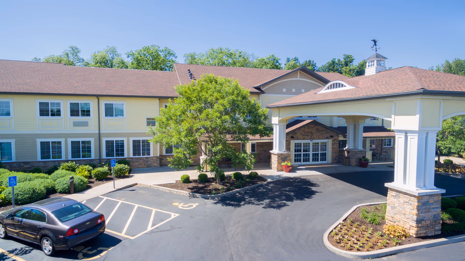 Front exterior view of Westview at Ellisville Assisted Living facility showing a two-story building with beige and stone facade, a covered entrance with white columns, a tree and landscaped bushes, and a parking area with handicap parking spaces and a parked car.