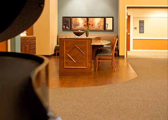 Interior view of a senior living facility showing a small dining area with a wooden table, chairs, a bench with patterned upholstery, a bowl of fruit, and a vase with flowers. The background includes a hallway with beige and orange walls and a framed picture above the bench.
