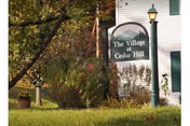 Outdoor view of a sign that reads 'The Village at Cedar Hill' situated on a grassy area with trees and bushes nearby, next to a white building with green shutters and a lamppost.
