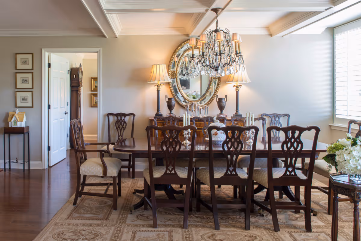 A traditional dining room with a large wooden dining table surrounded by eight matching chairs with cushioned seats. The room features a decorative chandelier hanging above the table, two table lamps on a sideboard against the wall, and a large round mirror between the lamps. The floor has a patterned area rug, and there is a window with white shutters letting in natural light. A small side table with a floral arrangement is visible on the right side.