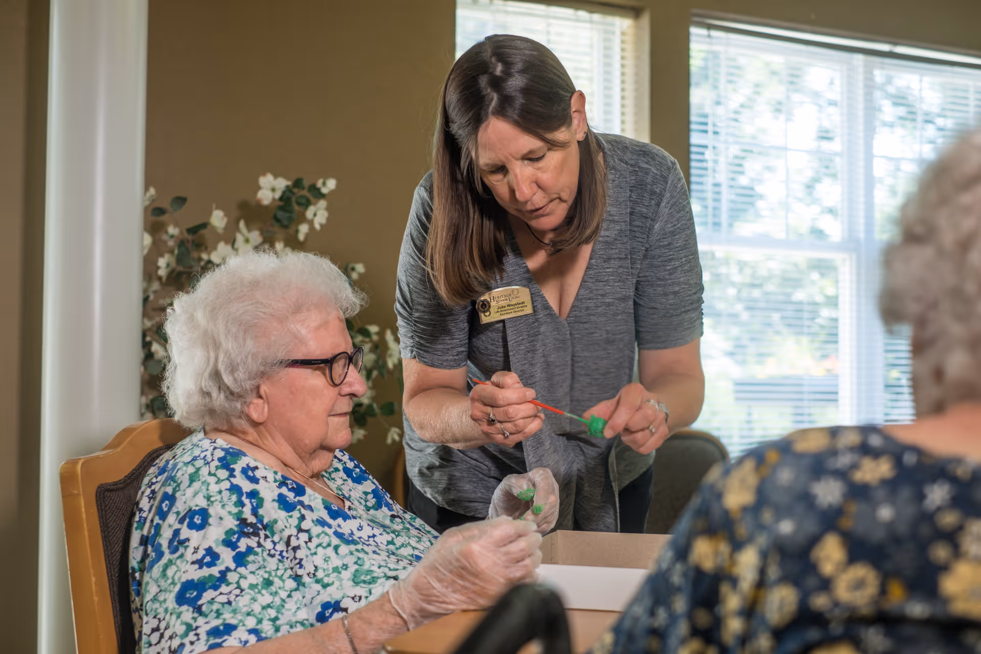 An elderly woman wearing glasses and a floral blouse is seated at a table, engaging in a craft activity with the assistance of a caregiver who is holding a paintbrush and helping her paint a small object green. Another elderly person is partially visible in the foreground. The setting is a well-lit room with large windows and a plant in the background.