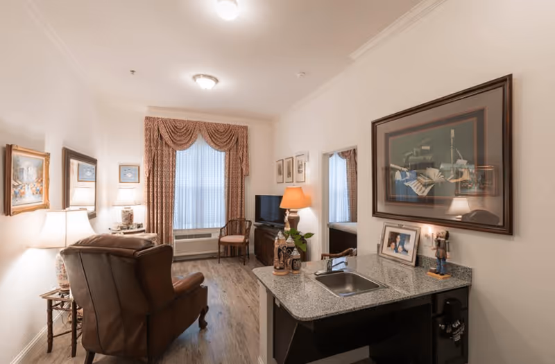 A cozy living room area with a brown leather armchair, two side tables with lamps, framed artwork on the walls, a window with patterned curtains, a TV on a stand, and a small kitchen counter with a sink and decorative items. A doorway leads to a bedroom with a bed visible.