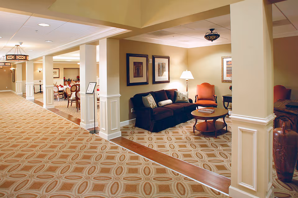 Interior view of a senior living facility showing a hallway with patterned carpet and a seating area. The seating area includes a dark sofa with cushions, an orange armchair, a round wooden coffee table, a floor lamp, and framed artwork on the walls. In the background, there is a dining area with tables and chairs set for a meal.