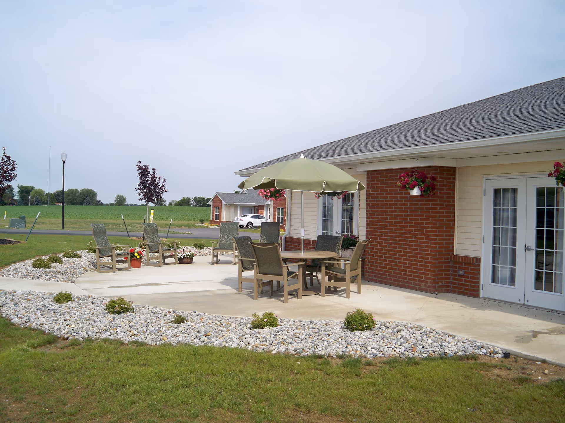Outdoor patio area at The Willows at Bellevue featuring a concrete seating area with a round table, six chairs, and a green umbrella. The patio is surrounded by landscaping with white rocks and small plants. The building has beige siding with a brick accent wall and glass double doors. In the background, there is a grassy field, a road, and another building with a parked white car.
