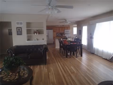 A spacious living and dining area with hardwood floors, featuring a dark leather sofa on the left, a round glass-top table with a plant in the foreground, a wooden dining table with chairs in the center, and a kitchen with wooden cabinets and white appliances in the background. Large windows with sheer curtains allow natural light to fill the room.