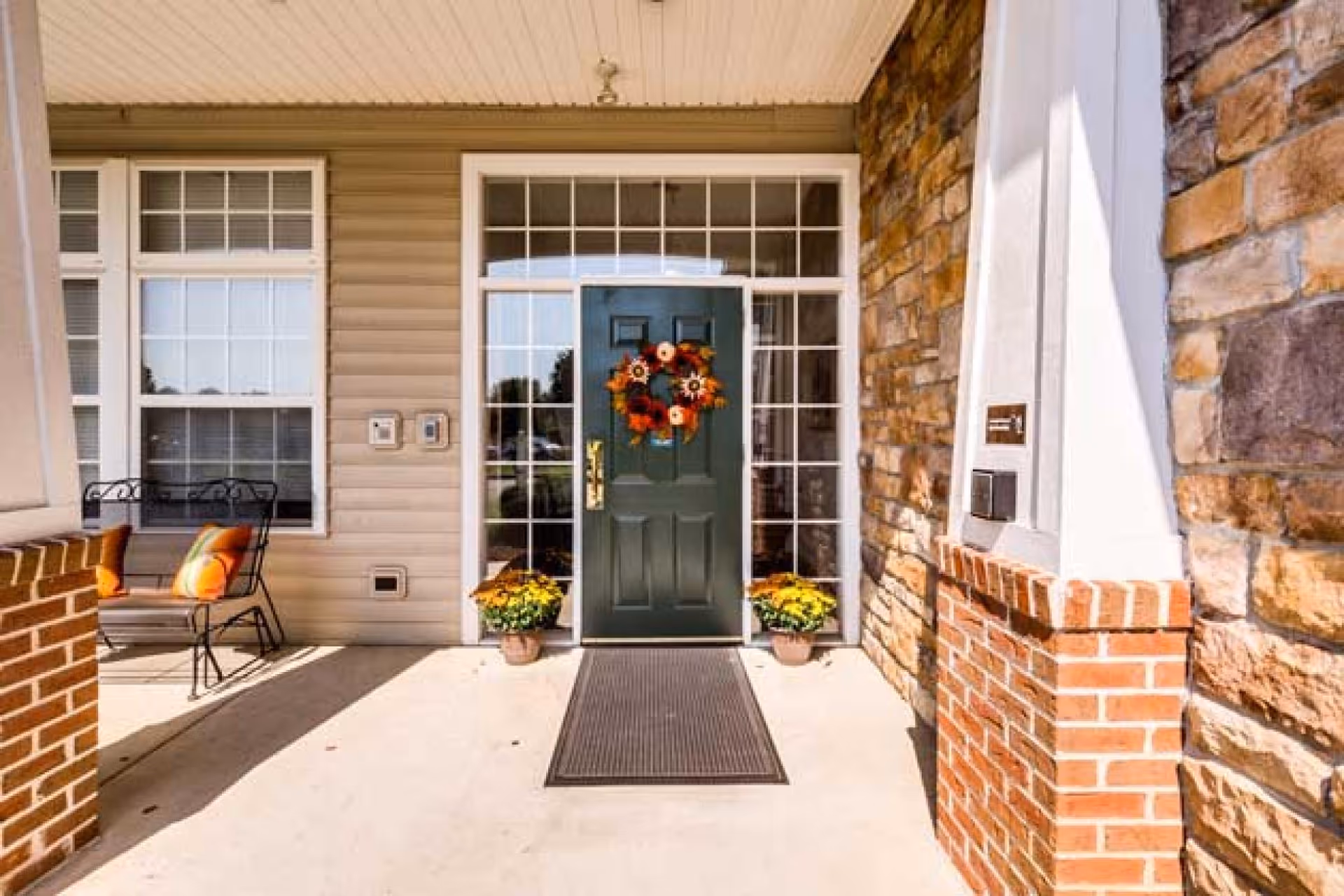 Front entrance of a building with a dark green door decorated with a fall-themed wreath. The door is flanked by large windows with white frames. Two potted yellow flowers are placed on either side of the door. There is a black doormat in front of the door. To the left, there is a seating area with a black metal chair and orange cushions. The exterior walls are a combination of beige siding and stone with brick accents.