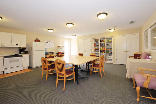 A spacious room with a kitchen area including a stove, refrigerator, and microwave on the left side. In the center, there are two square tables pushed together with wooden chairs around them. On the right side, there is a table covered with a cloth and a purple cushioned chair. The back wall has shelves stocked with various items and two closed double doors. The room is well-lit with ceiling lights and has a carpeted floor.