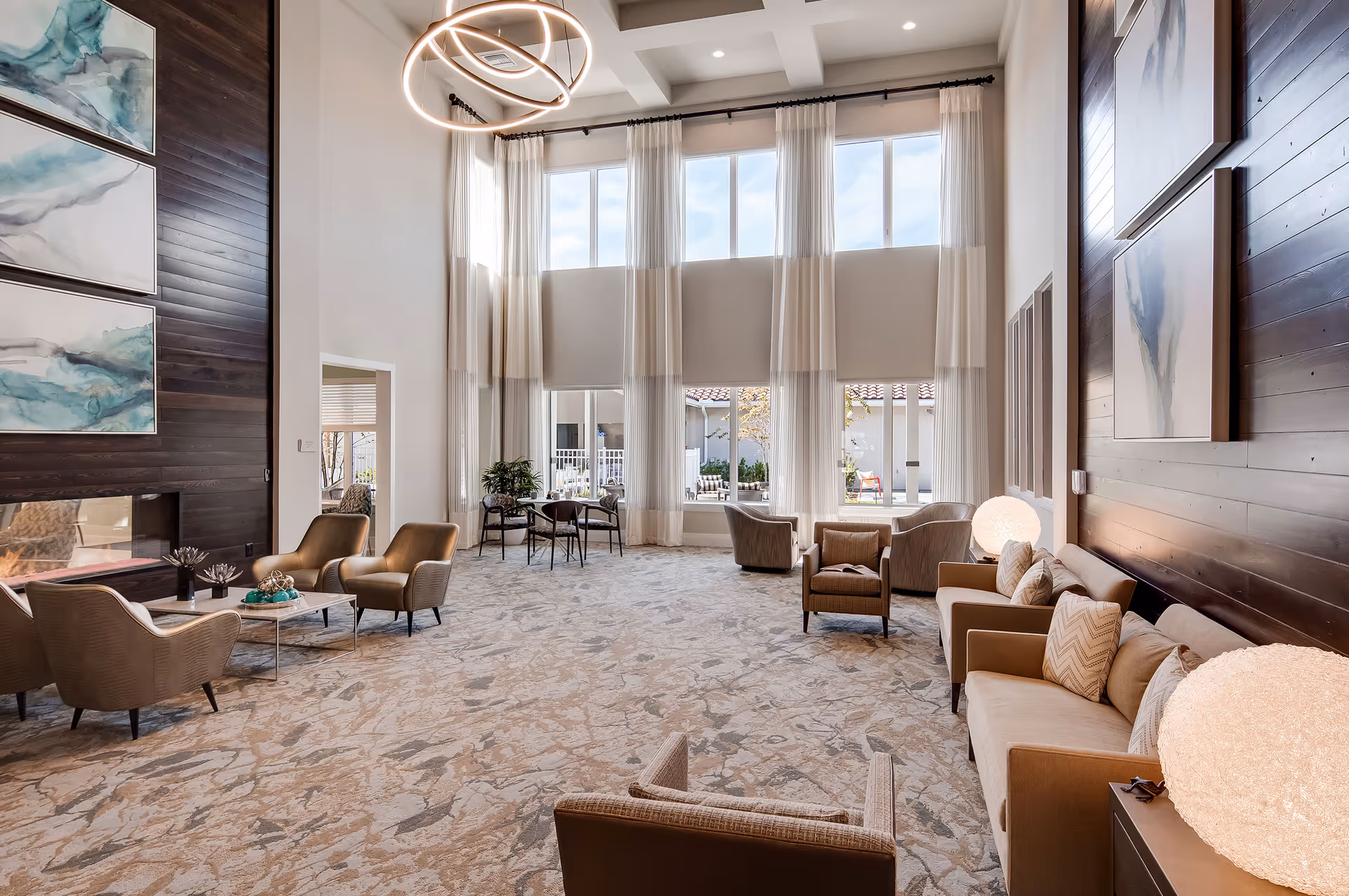 A spacious and well-lit living room area with high ceilings and large windows covered with sheer curtains. The room features multiple comfortable armchairs and sofas arranged around small tables, a modern circular chandelier, abstract artwork on dark wood-paneled walls, and two glowing spherical lamps on side tables.