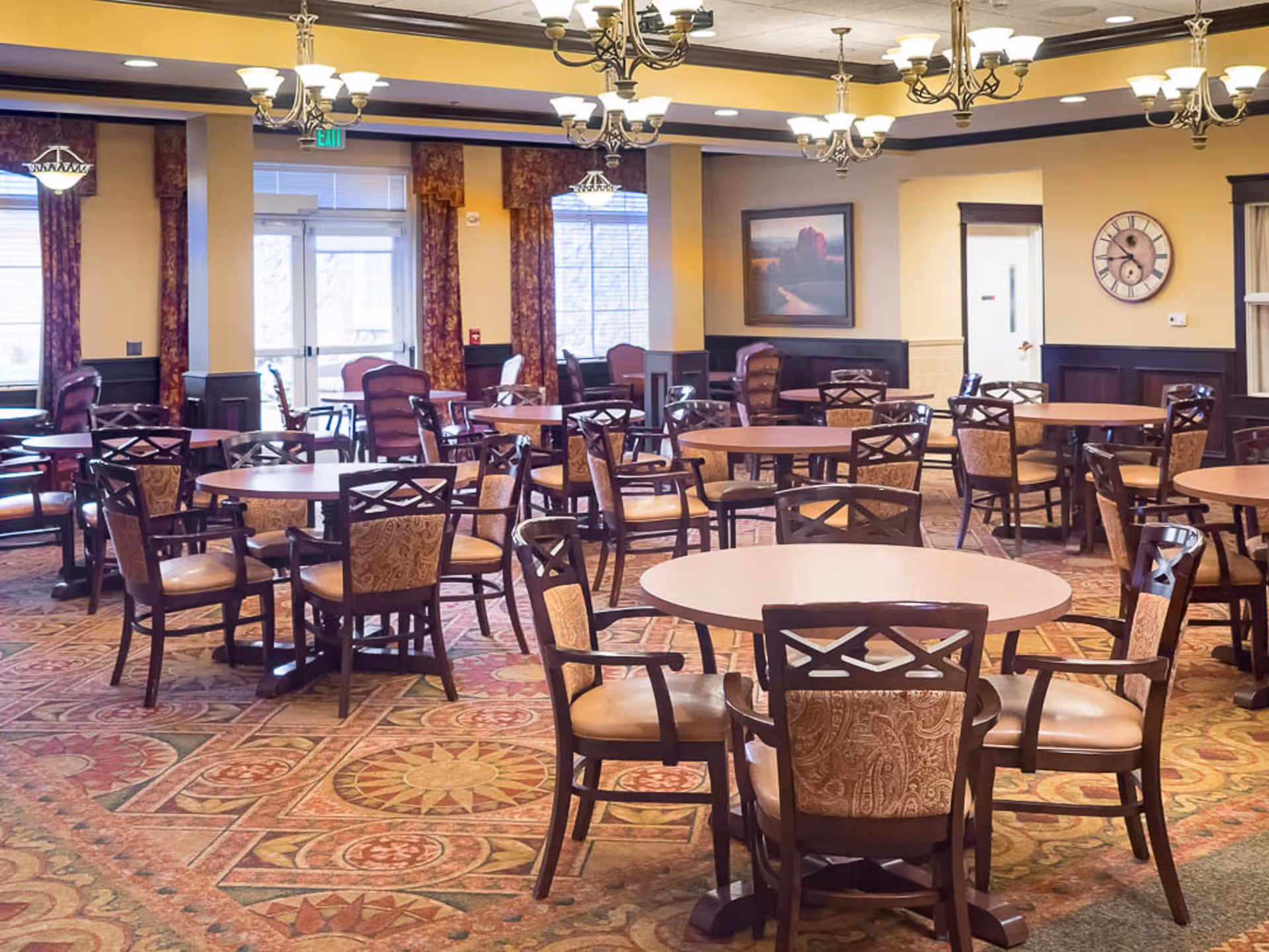 A spacious dining room with multiple round wooden tables surrounded by cushioned chairs with patterned upholstery. The room features warm yellow walls, large windows with decorative curtains, chandeliers hanging from the ceiling, a large wall clock, and a framed landscape painting. The floor is covered with a patterned carpet.