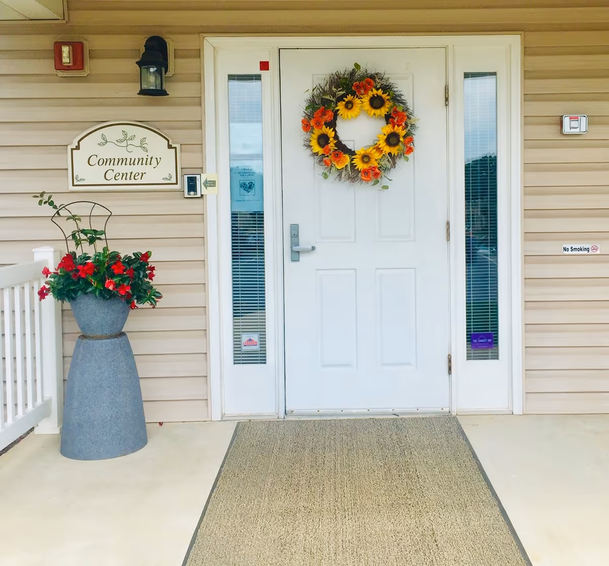 Front entrance of a beige-sided community center with a white door decorated with a sunflower wreath, flanked by narrow windows and a planter of red flowers.