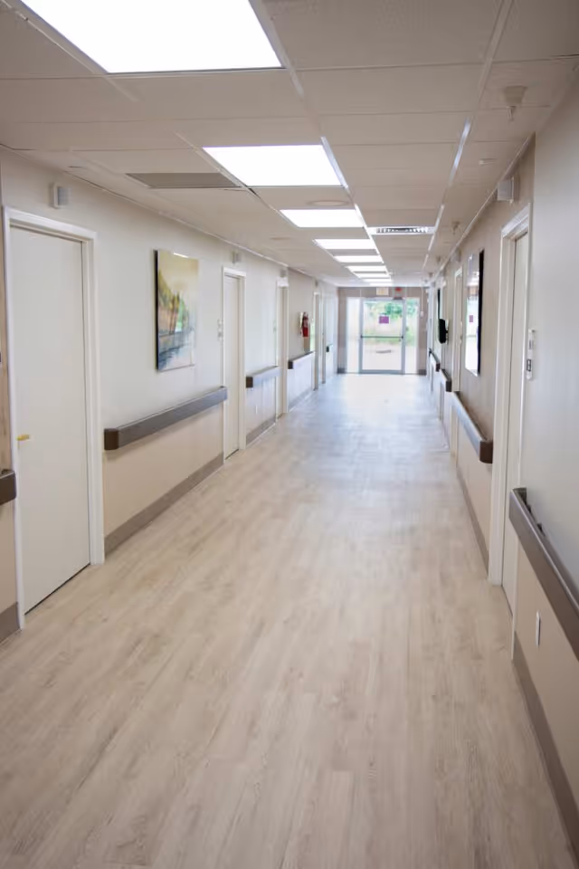 A long, clean hallway in a healthcare facility with light wood flooring, white walls, multiple closed doors on both sides, handrails along the walls, ceiling lights, and a glass exit door at the far end.