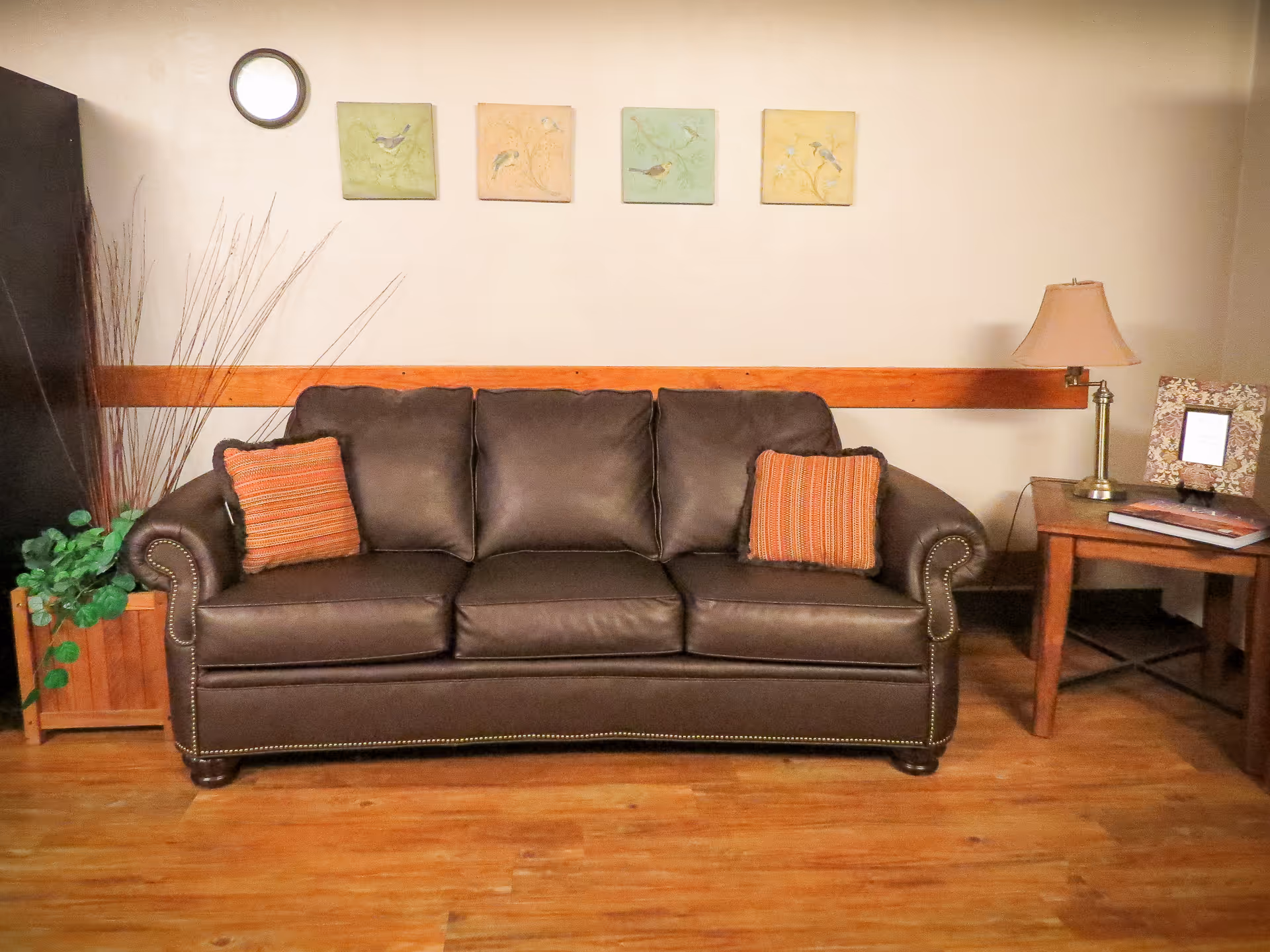 A cozy living room area featuring a dark brown leather sofa with two orange striped pillows. Behind the sofa, there are four small square paintings of birds on the wall. To the right of the sofa is a wooden side table with a lamp and a framed photo. To the left of the sofa is a wooden planter with green plants and tall decorative sticks. The floor is wooden and the walls are light-colored with a wooden trim.