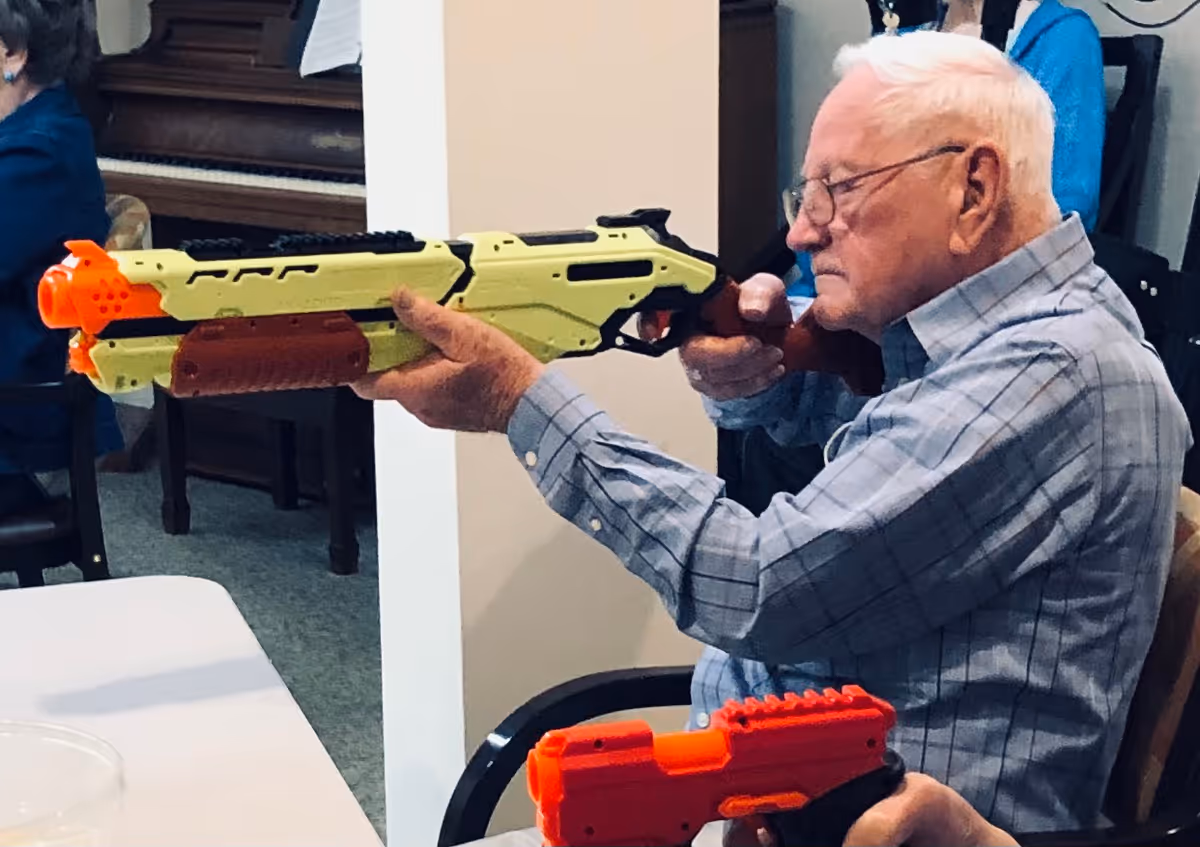 An elderly man wearing glasses and a checkered shirt is sitting indoors aiming a large toy gun that is yellow and orange. Another person nearby is holding a smaller orange toy gun. There is a piano and other furniture in the background.