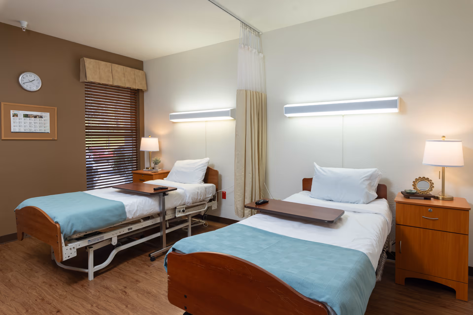 A clean and well-lit room with two hospital-style beds, each with white sheets, pillows, and light blue blankets. Each bed has a wooden bedside table with a lamp and some decorative items. The room has wooden flooring, a window with brown blinds and a beige valance, a wall clock, and a framed picture. A privacy curtain is partially visible between the beds.