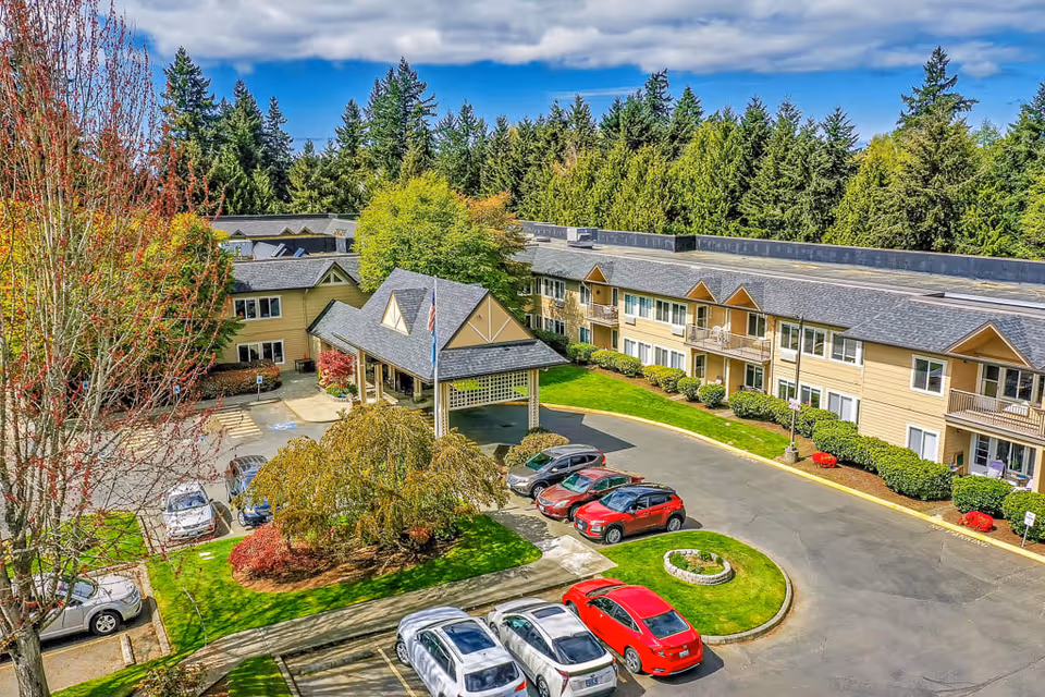 Aerial view of Solstice Senior Living At Normandy Park showing a two-story building with beige siding and dark gray roofs surrounded by trees and greenery. There is a covered entrance with an American flag, a circular driveway with parked cars, and well-maintained landscaping including bushes and a small garden area.
