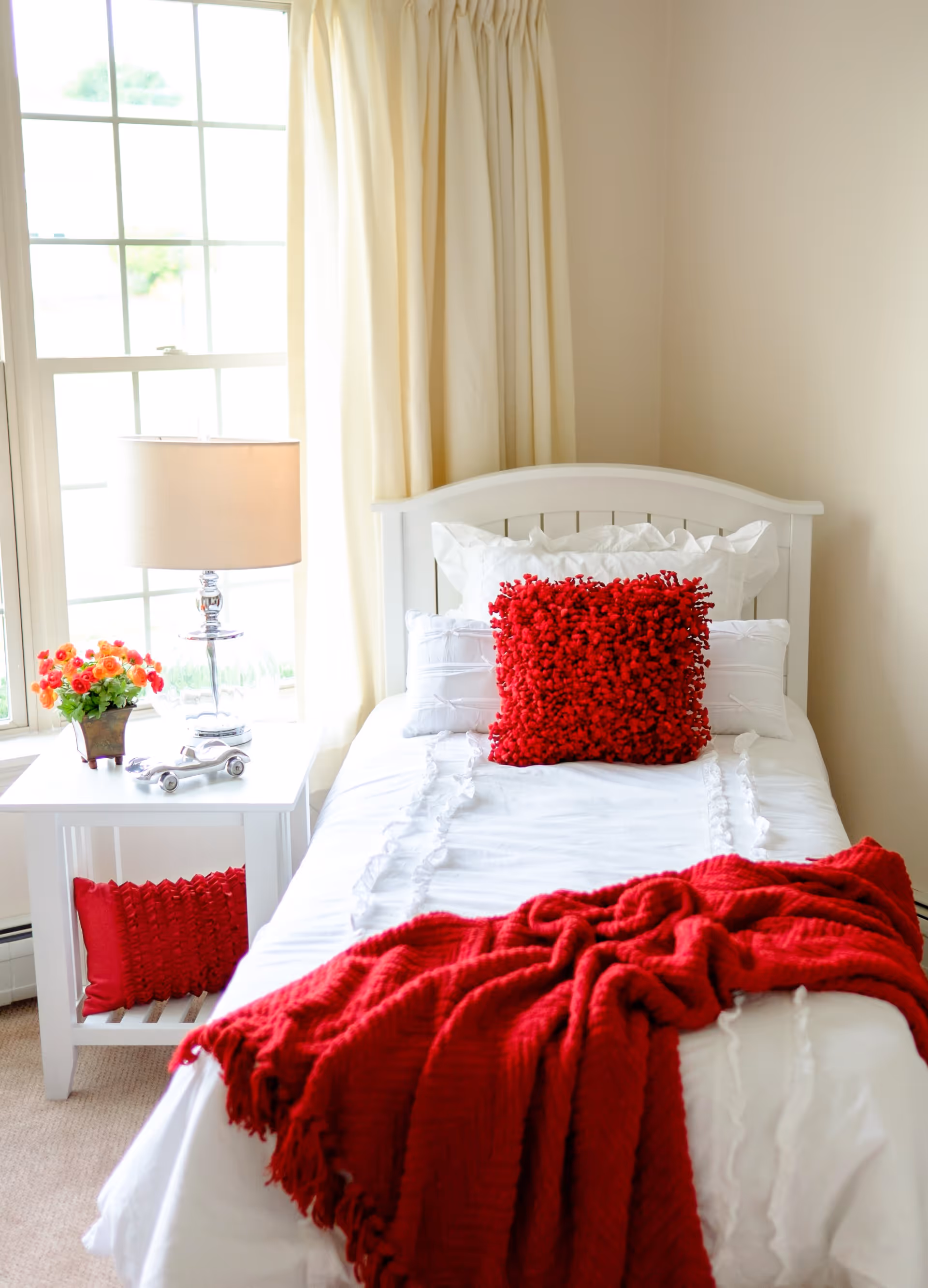 A bright bedroom with a white single bed featuring white bedding, a textured red pillow, and a red throw blanket. Next to the bed is a white nightstand with a clear glass lamp, a small decorative silver car, and a pot of orange flowers. A window with cream-colored curtains is behind the nightstand.