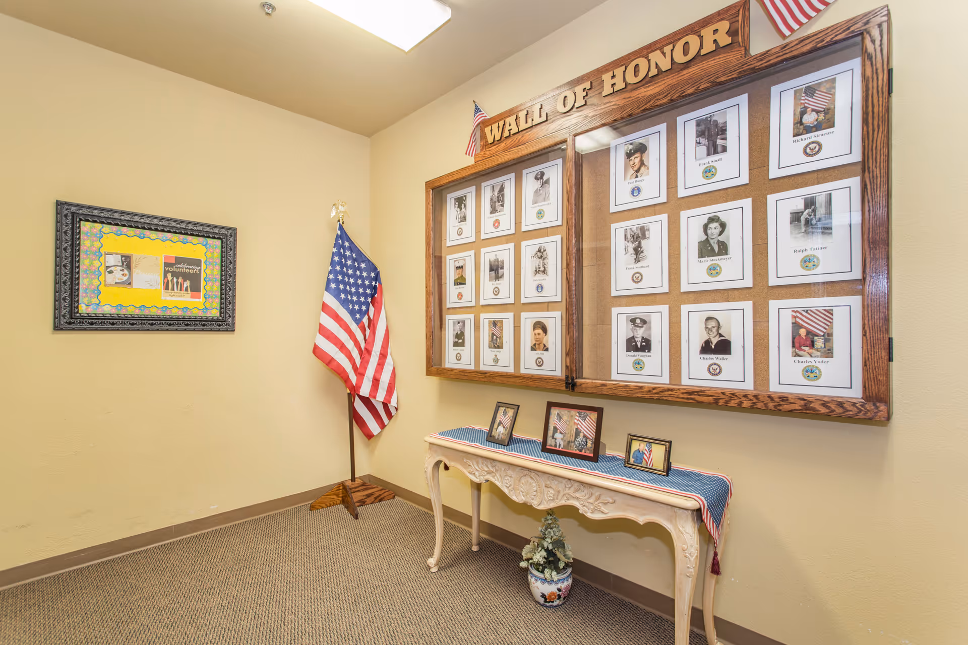 Interior corner of a senior living facility with a Wall of Honor display featuring framed photos of veterans, an American flag on a stand, a decorative table with small framed pictures and a potted plant underneath, and a bulletin board on the adjacent wall.