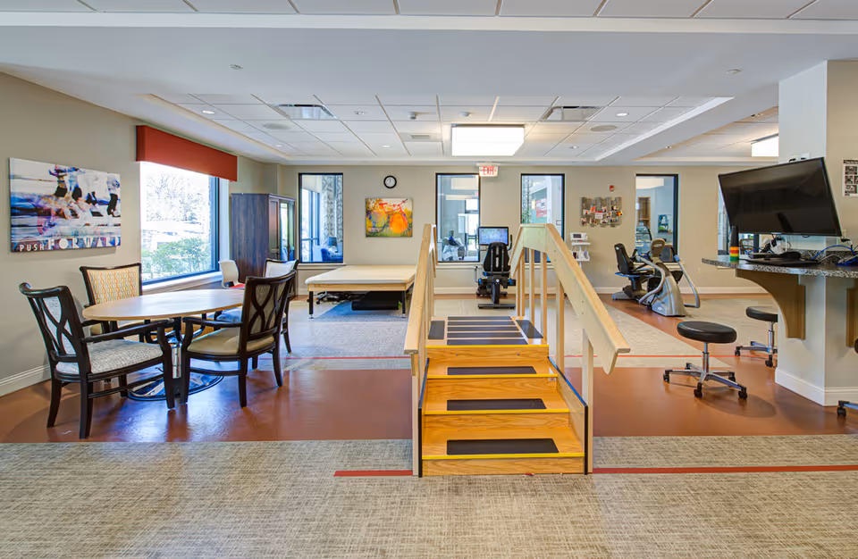 A rehabilitation room featuring a wooden staircase with handrails in the center, exercise equipment including stationary bikes and a recumbent bike, a table with four chairs on the left, and a computer workstation with stools on the right. The room has large windows, colorful artwork on the walls, and a ceiling with recessed lighting.