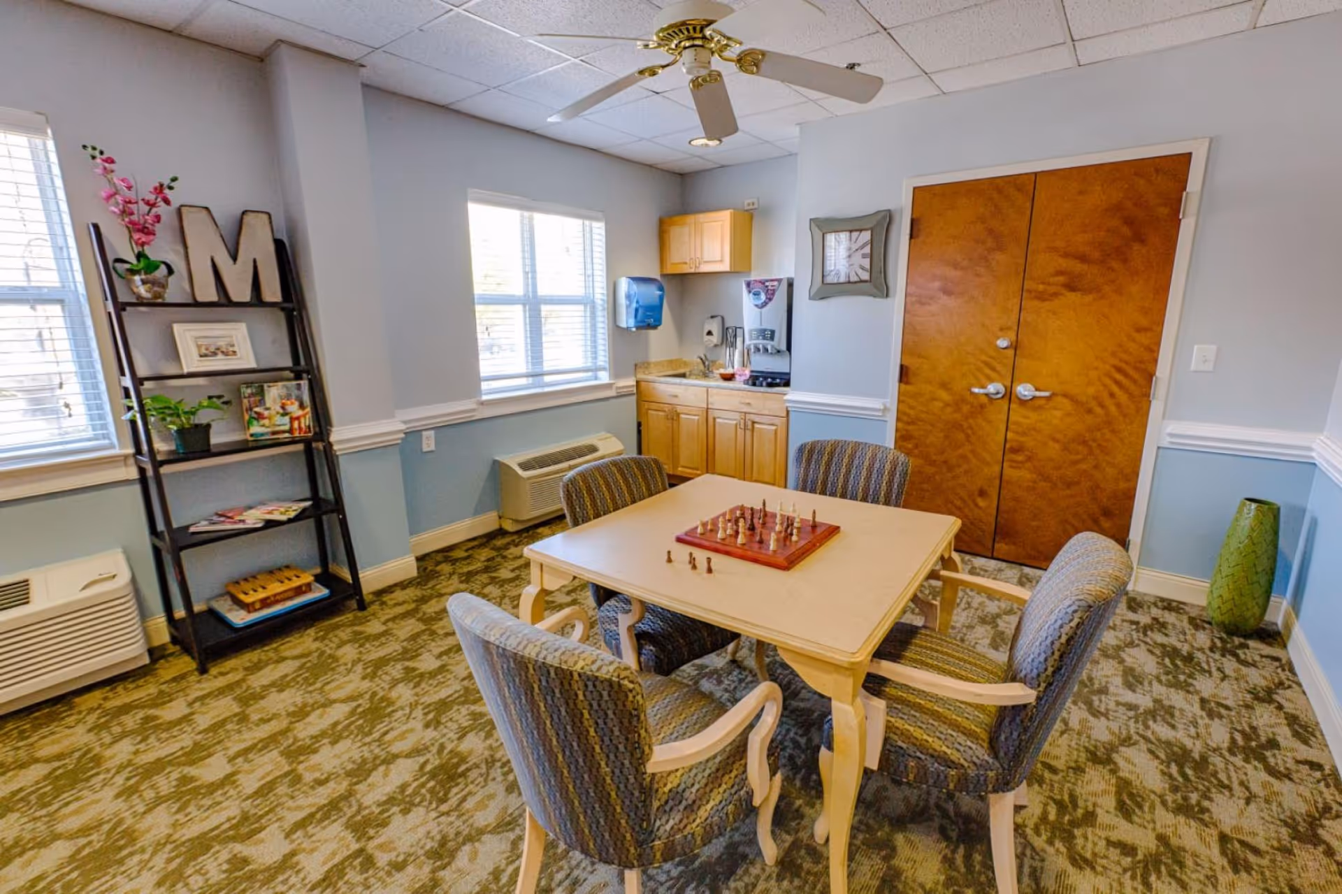 Bright common room with a square table and four upholstered chairs around a chessboard, kitchenette cabinets, bookshelf and double doors.