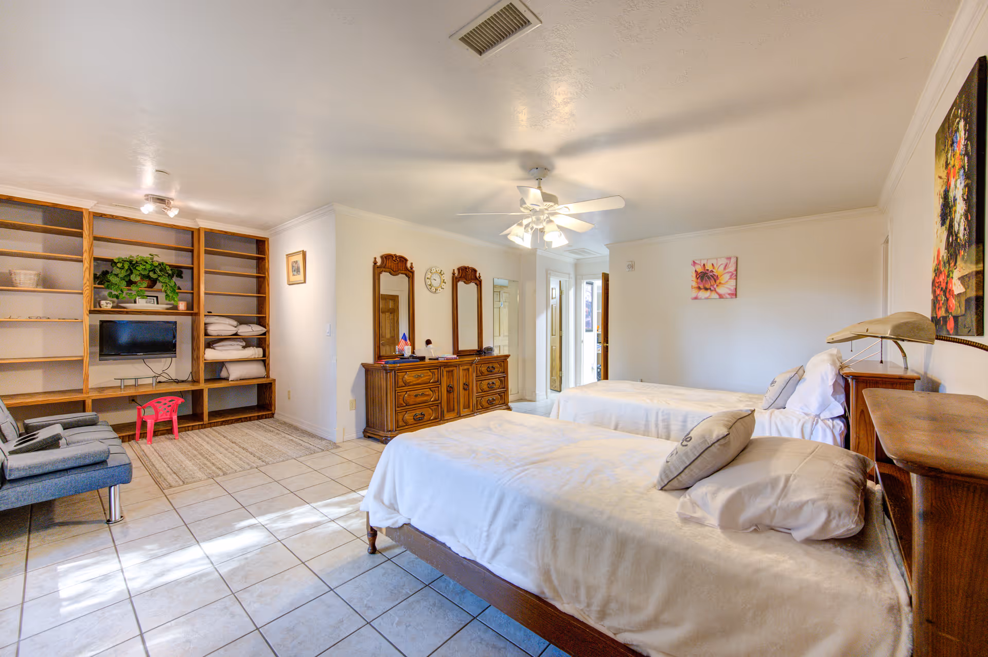 A bright senior care bedroom with two single beds covered in white linens, a wooden dresser with two mirrors and a clock above it, a built-in wooden shelving unit with a small TV and some folded linens, two blue chairs, a small pink stool, and tiled flooring. The room has white walls and ceiling with a ceiling fan and light fixture.