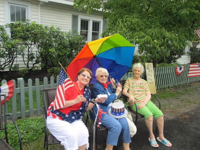 Three elderly women sitting outdoors on chairs near a green picket fence. One woman is holding a rainbow-colored umbrella and an American flag, another is holding a small American flag and wearing a patriotic hat, and the third woman is sitting casually. There are patriotic decorations on the fence and a sign that reads '4th of July BBQ'.