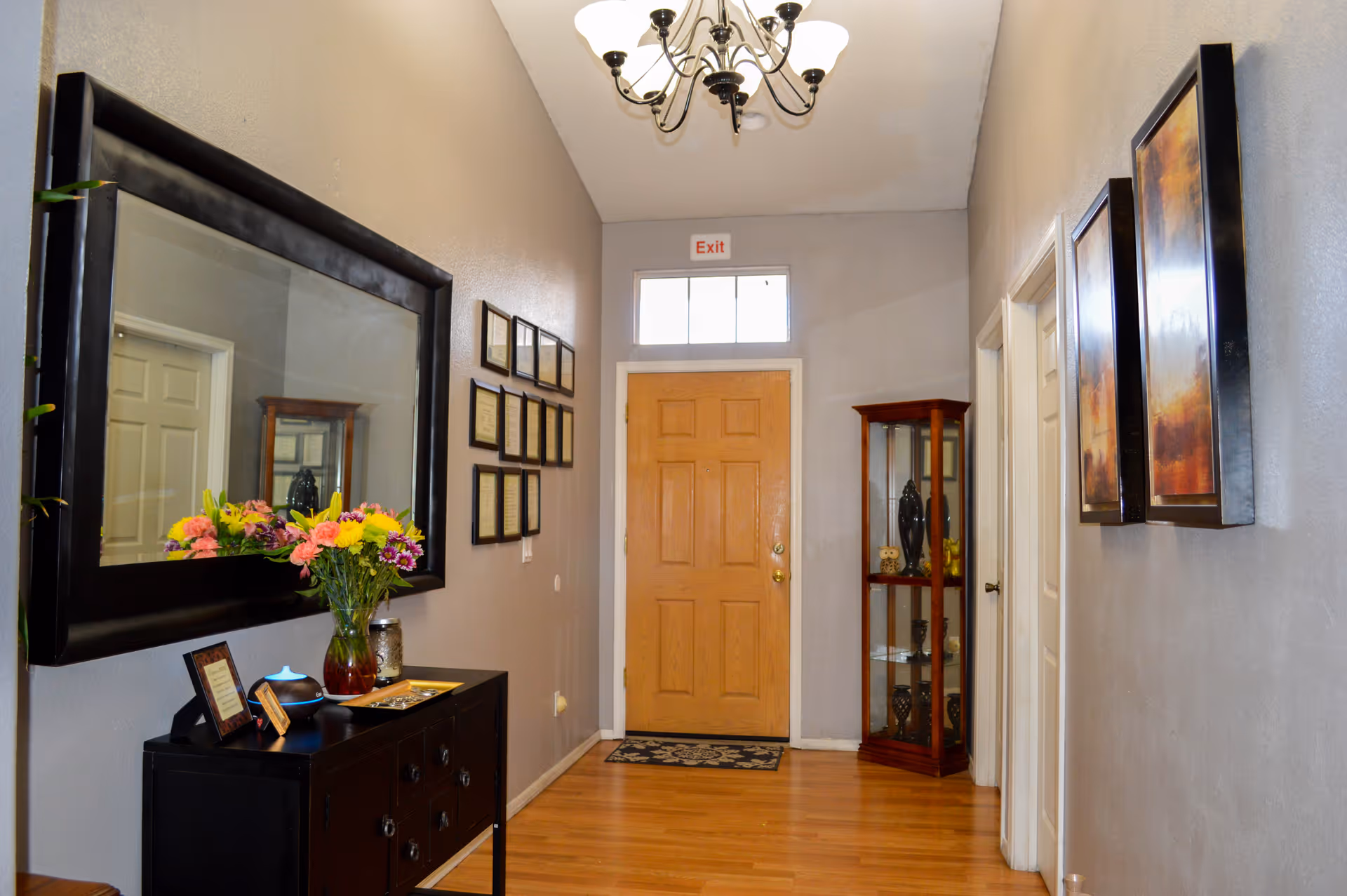 Interior hallway of La Bella Vita Assisted Living Home with a wooden front door, a large mirror on the left wall above a black console table with a vase of colorful flowers, framed certificates on the left wall, a glass display cabinet with decorative items on the right, and two framed paintings on the right wall. A chandelier hangs from the ceiling.