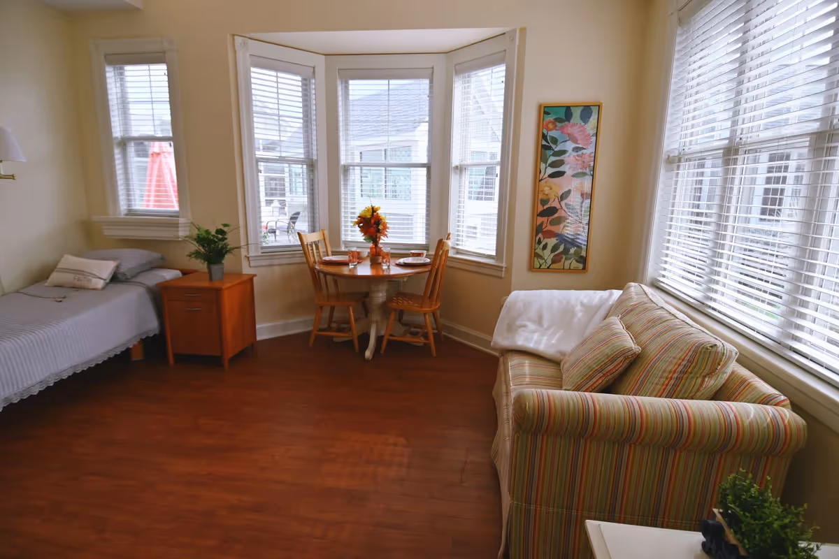 Sunny studio-style living area with a single bed, small round dining table by bay windows, and a striped sofa.